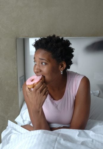 Woman in pink shirt taking a bite of a pink frosted doughnut (donut) while sitting up in bed. (iStockPhoto NYC 9/13/05)