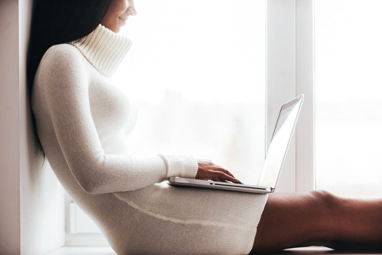 Cropped image of attractive young African woman in warm sweater working on laptop while sitting on the window sill